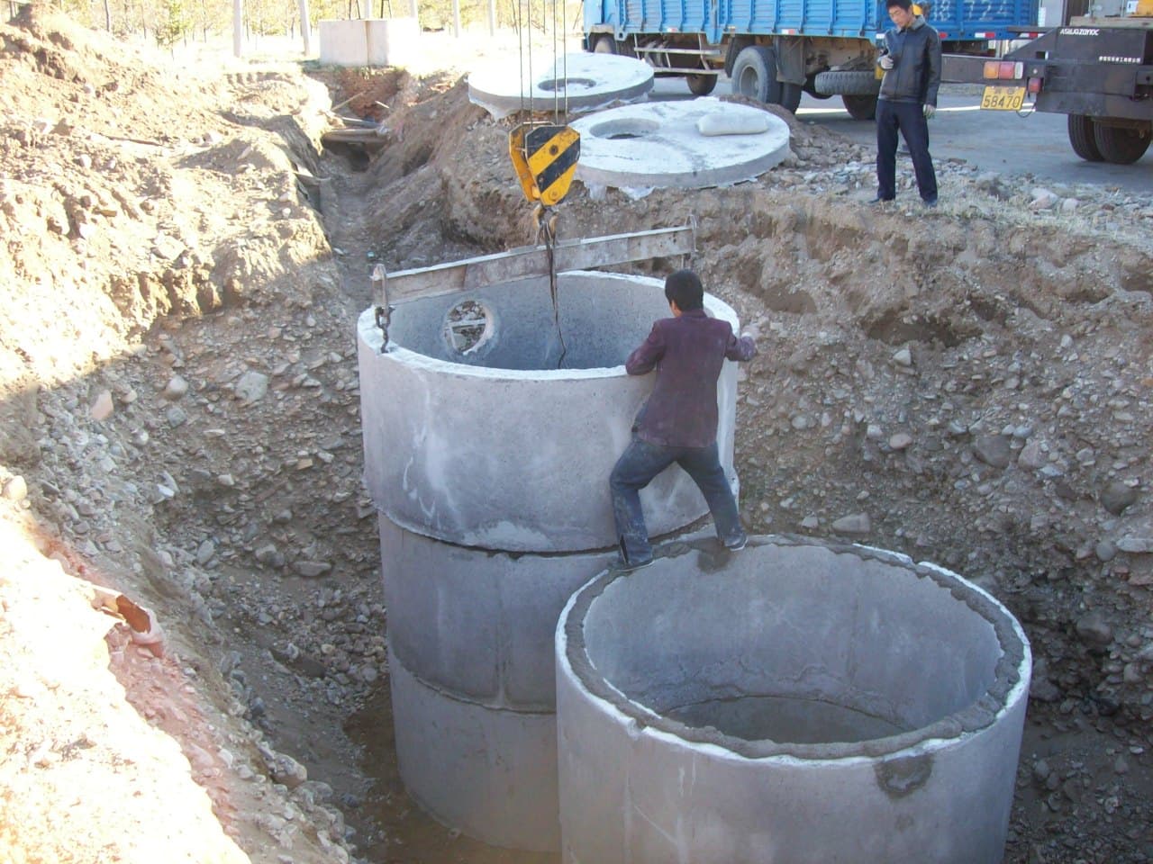 A crane lowering precast concrete septic tank sections into an excavated pit while a worker guides them into place, with more concrete lids on a delivery truck behind