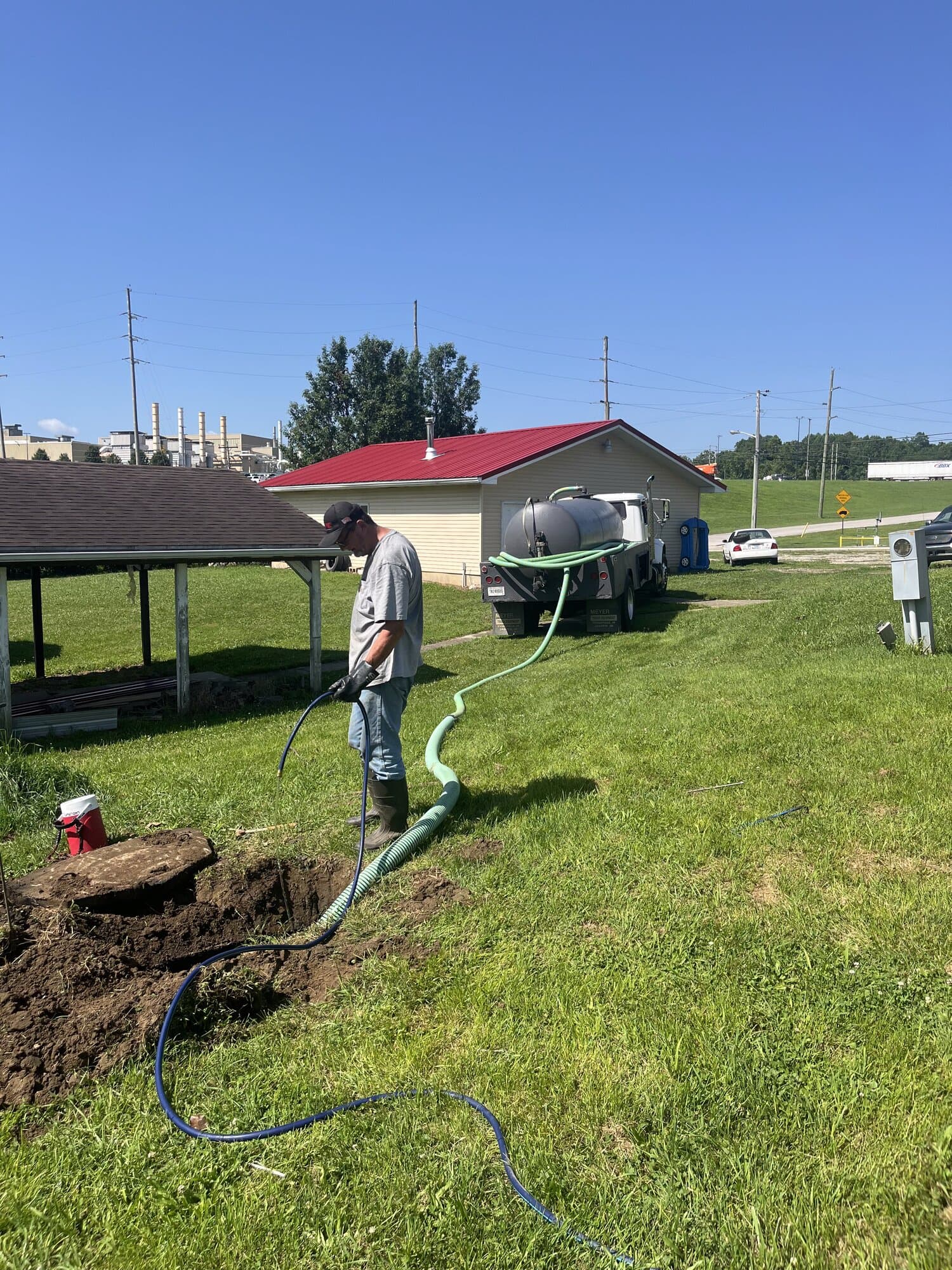 A Quick Pump and Clean technician standing over a freshly hand-dug hole with the septic tank lid uncovered and set aside, with the pump truck parked by the house behind him