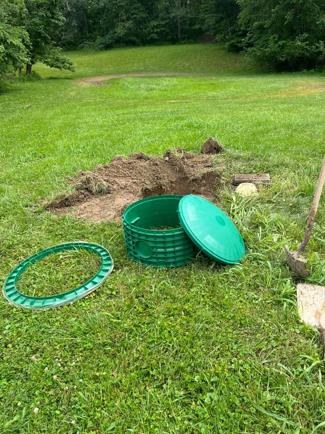 A green plastic septic riser extension ring and lid staged next to an excavated access hole before installation