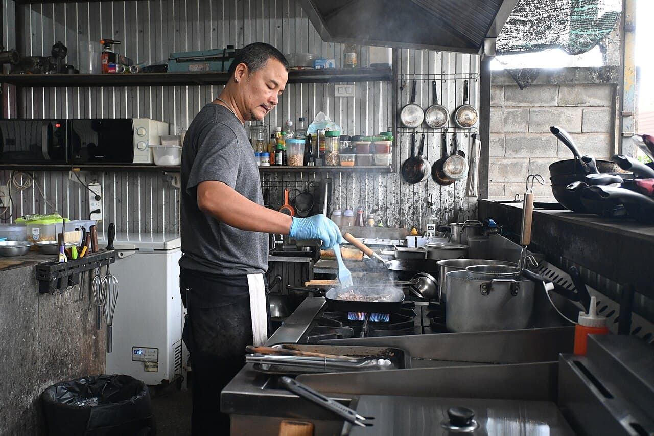 A cook working a hot pan over an open flame in a busy commercial kitchen, the fry station and squeeze bottles beside him, where fats, oils, and grease first head for the drain