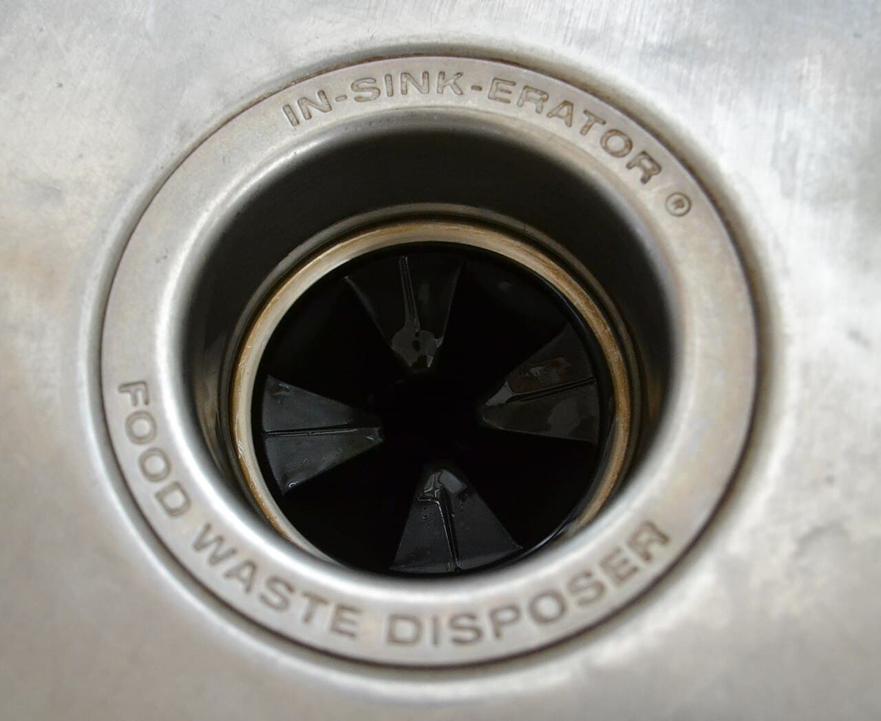 Top-down view into a stainless steel kitchen sink garbage disposal, with the grinding chamber and splash guard visible and the words food waste disposer stamped around the rim