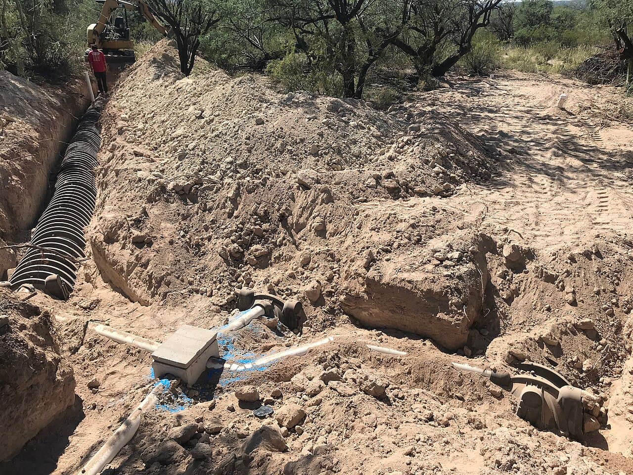 A septic drain field under construction on a rural lot, with an open trench of perforated chamber pipe, a distribution box, and an excavator working the sandy soil
